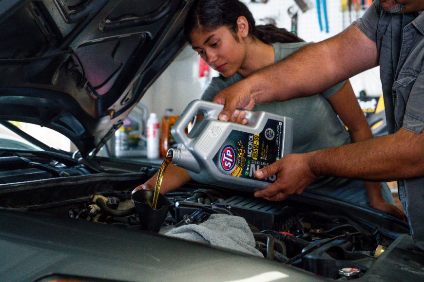 A daughter watches as a her father pours STP synthetic motor oil to show her how to change the car's oil.